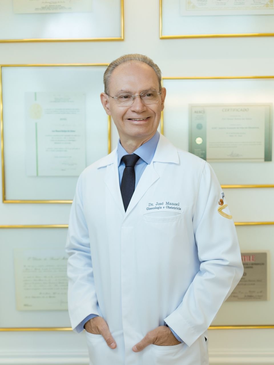 Dr. José Manoel de jaleco branco, diante da parede com seus certificados